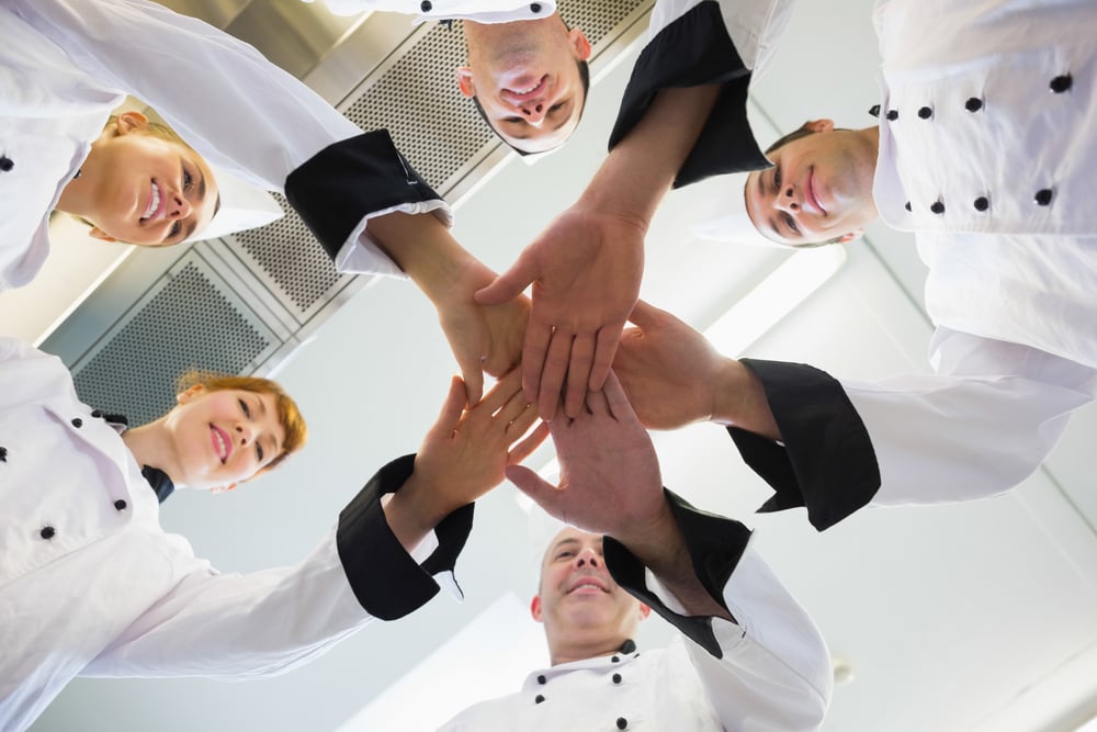 Chefs joining hands in a circle wearing uniforms in a kitchen